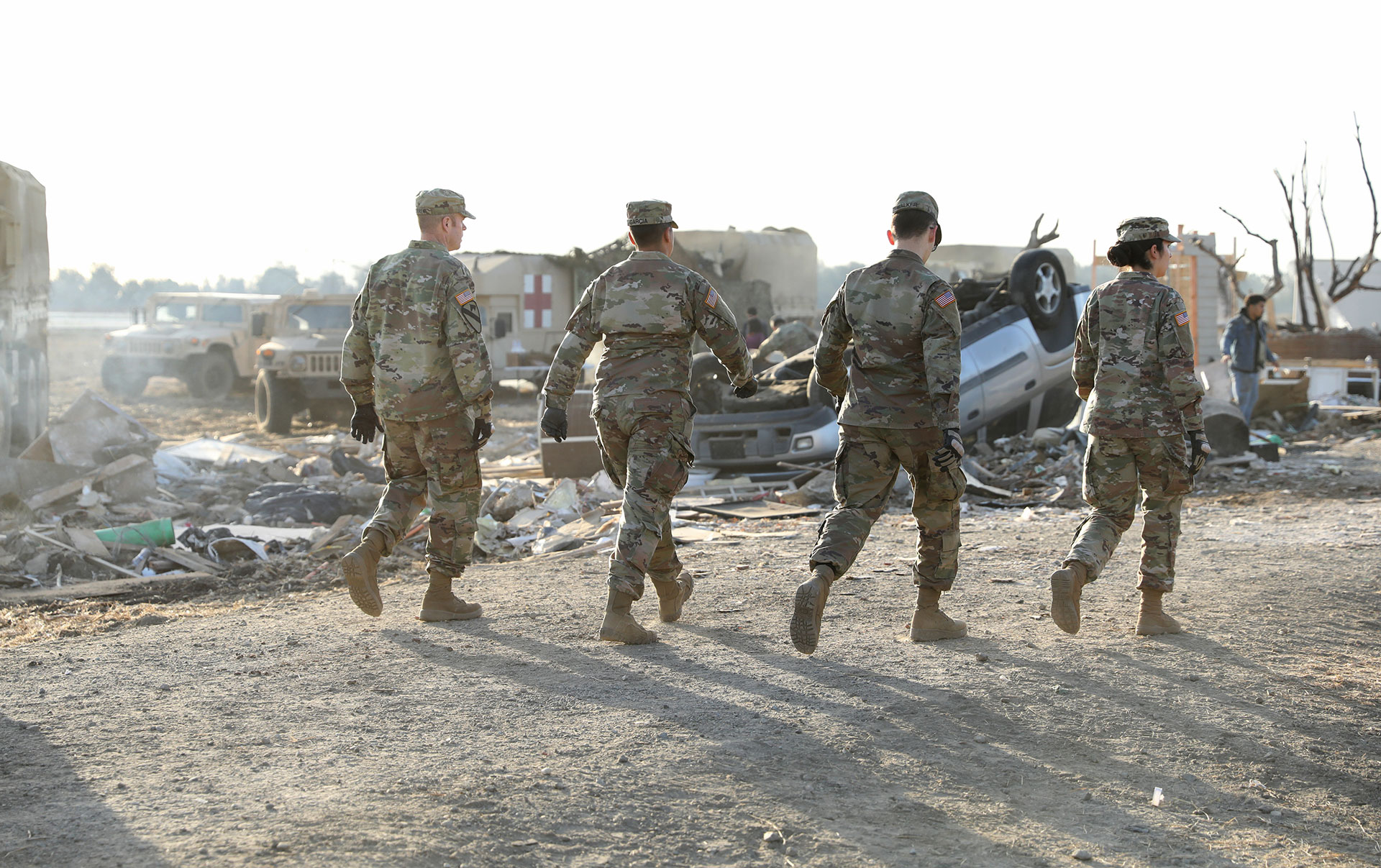 Four Soldiers walking through disaster scene with emergency vehicles parked in the background